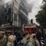 Thick smoke billowing from the top floors of the Flourish Stay B&B building during the tragic Malviya Nagar hotel fire in Delhi as fire services operate in a congested market lane.